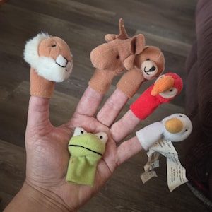 May include: A hand displaying five finger puppets: a lion, a bear, a bird, a frog, and a duck. The puppets are made of fabric and have detailed facial features. The lion has a fluffy mane, the bear has a black nose, and the bird has a red beak.