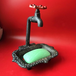 May include: A black cast iron soap dish shaped like a faucet, with a red handle and a green bar of soap. The dish has an ornate, textured design and sits on a red background.