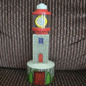 May include: A toy lighthouse with a red top and a clear, yellow-accented light section. The body is gray, resembling stone, with a small blue window and a red door. The base has a door and green foliage.