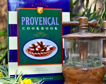May include: A wooden spice grinder with a glass jar filled with dried herbs. The grinder is sitting on a wooden table next to a cookbook titled "A Little Provençal Cookbook" by Lynn Bright. There are also some yellow flowers and purple lavender flowers on the table.