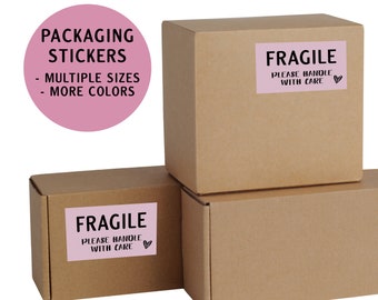 May include: Three brown cardboard boxes stacked on top of each other. The top box has a pink "Fragile" sticker with the text "Please handle with care" and a heart. The bottom box also has a pink "Fragile" sticker with the text "Please handle with care" and a heart. A pink circle with the text "Packaging Stickers" and "Multiple Sizes" and "More Colors" is in the top left corner of the image.