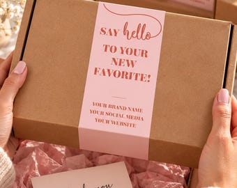 May include: A brown cardboard shipping box wrapped with a light pink paper belly band featuring the text Say hello TO YOUR NEW FAVORITE! and placeholders for brand details. Inside, pink tissue paper holds a Thank you card with a heart icon.