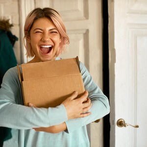 May include: A person with pink hair is holding a brown cardboard box in front of them. They are smiling and looking at the camera.
