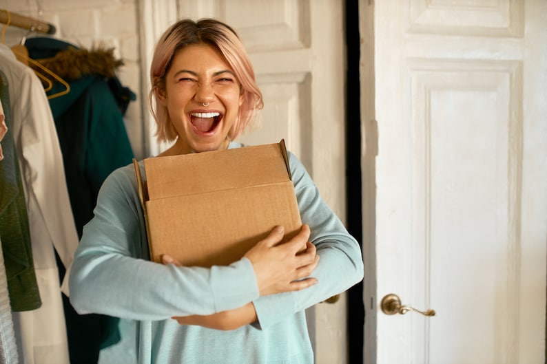 May include: A person with pink hair is holding a brown cardboard box in front of them. They are smiling and looking at the camera.