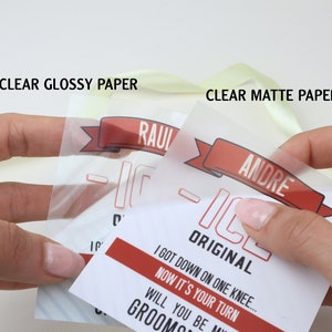 May include: Two transparent paper tags with red and black text and graphics. One tag is labeled CLEAR GLOSSY PAPER and features the name RAUL, while the other is labeled CLEAR MATTE PAPER and features the name ANDRE, with the text I GOT DOWN ON ONE KNEE... NOW IT'S YOUR TURN WILL YOU BE MY GROOMSMAN.
