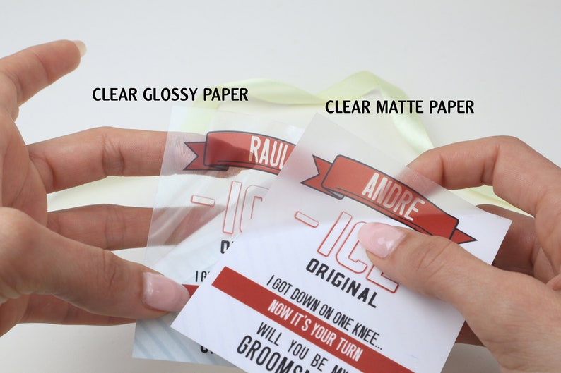 May include: Two transparent paper tags with red and black text and graphics. One tag is labeled CLEAR GLOSSY PAPER and features the name RAUL, while the other is labeled CLEAR MATTE PAPER and features the name ANDRE, with the text I GOT DOWN ON ONE KNEE... NOW IT'S YOUR TURN WILL YOU BE MY GROOMSMAN.