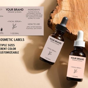 May include: Two brown glass serum bottles with dropper tops, each labeled "YOUR BRAND" and "FACIAL SERUM." The labels are light pink with a botanical illustration. Text on a separate label reads "CUSTOM COSMETIC LABELS."