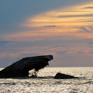 Puede incluir: Una gran estructura de hormigón, posiblemente un barco, parcialmente sumergida en el agua al atardecer. El cielo es una mezcla de rosa y azul con nubes.