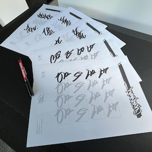 May include: A stack of white paper sheets with calligraphy practice. The sheets feature black and gray lettering, with a black pen on the side. The pen has red and white text on it. The calligraphy appears to be in a stylized script.