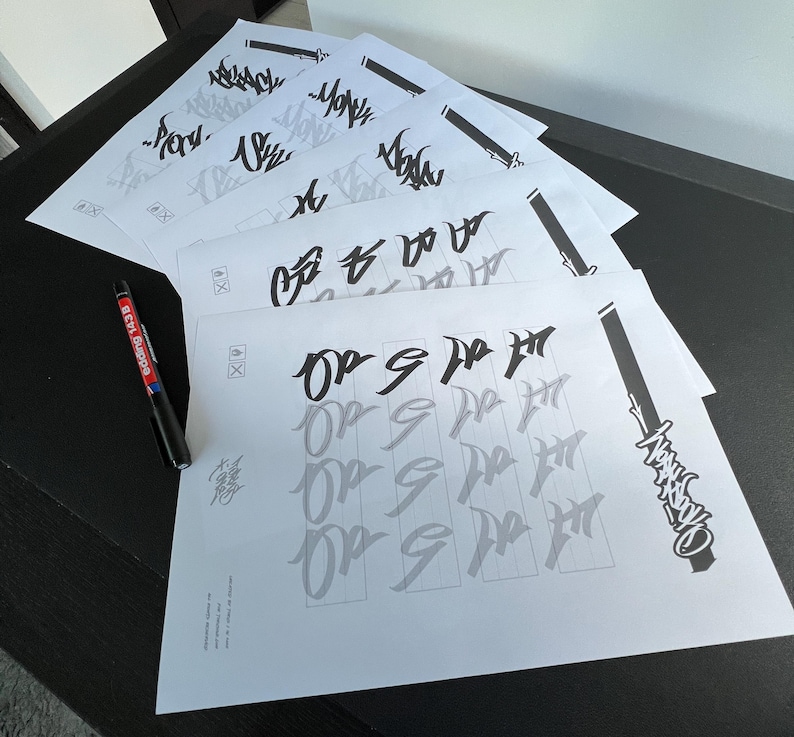 May include: A stack of white paper sheets with calligraphy practice. The sheets feature black and gray lettering, with a black pen on the side. The pen has red and white text on it. The calligraphy appears to be in a stylized script.