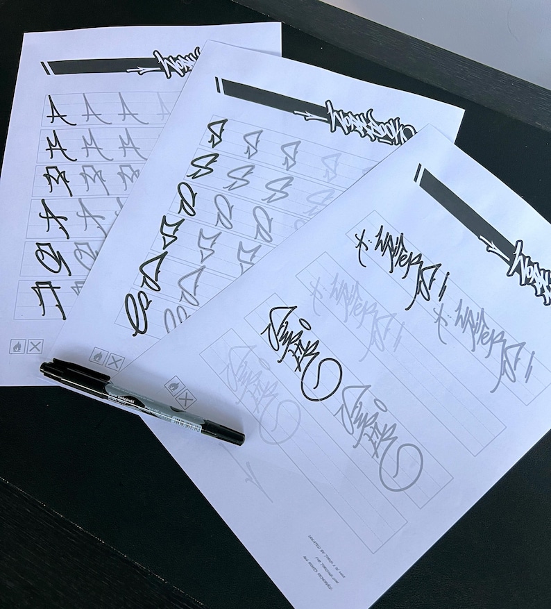 May include: Four sheets of white paper with black ink lettering and designs. The sheets feature various letter styles and a signature. A black marker pen rests on the bottom left corner of the papers. The papers are on a dark surface.