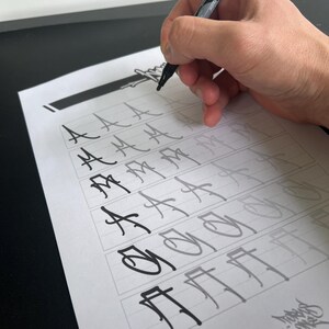 May include: A hand holding a black marker is writing on a sheet of paper with a grid of boxes. The paper contains stylized letter "A" and other characters in various stages of completion. The background is a black surface and a white wall.
