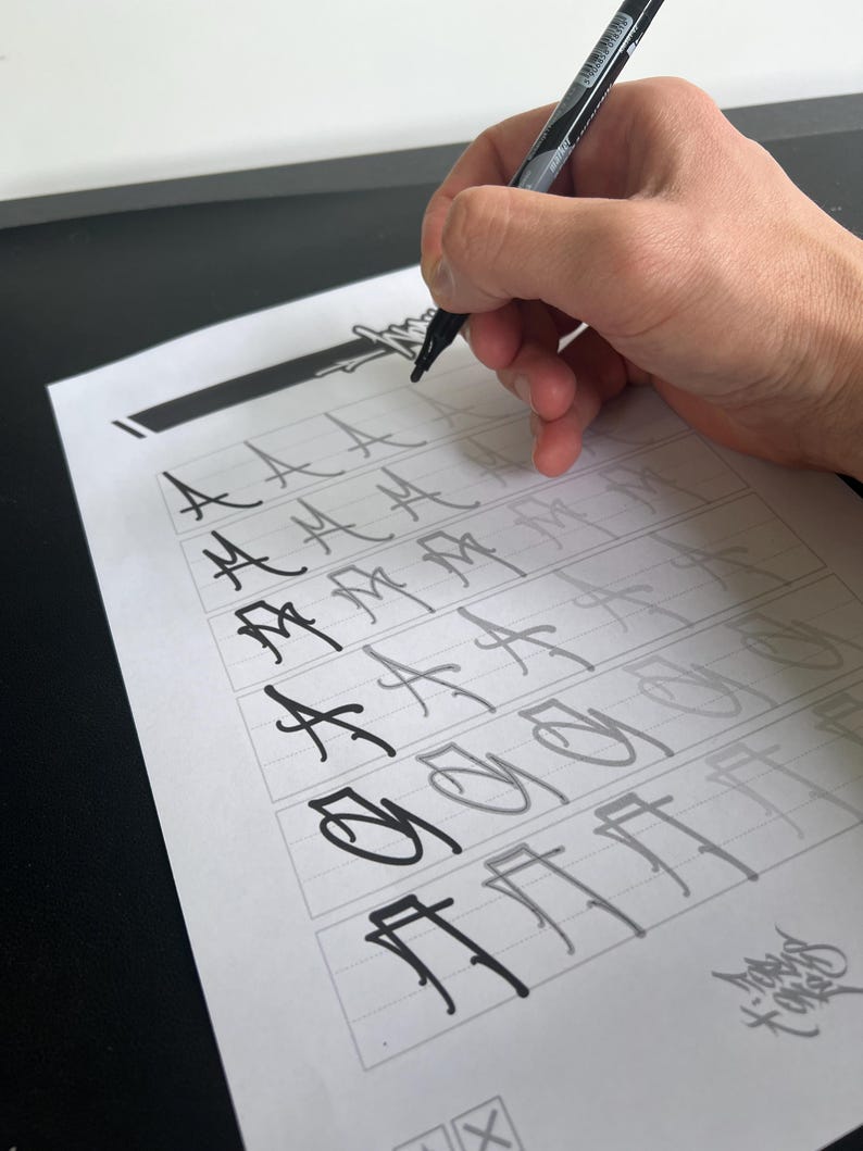 May include: A hand holding a black marker is writing on a sheet of paper with a grid of boxes. The paper contains stylized letter "A" and other characters in various stages of completion. The background is a black surface and a white wall.