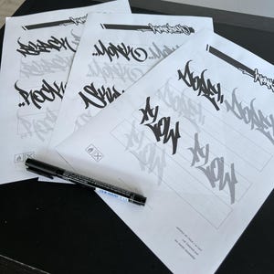 May include: Four white sheets of paper with black graffiti-style lettering and a black marker. The sheets have a black bar at the top and a faint shadow of the lettering. The marker is lying on top of the sheets.