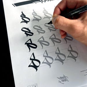 May include: A hand using a black marker to trace a stylized letter "S" on a white sheet of paper. The paper features multiple iterations of the letter in varying shades of gray and black, along with a decorative logo and other graphic elements.