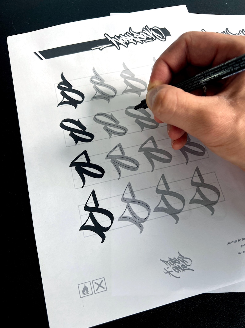 May include: A hand using a black marker to trace a stylized letter "S" on a white sheet of paper. The paper features multiple iterations of the letter in varying shades of gray and black, along with a decorative logo and other graphic elements.