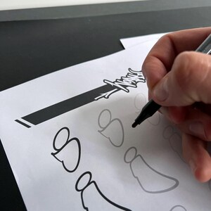 May include: A hand holding a black marker is drawing a black outline of a design on white paper. The design appears to be a stylized angel or heart shape. Other faint outlines of the design are visible.