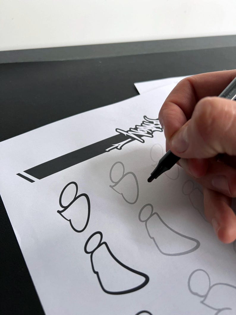 May include: A hand holding a black marker is drawing a black outline of a design on white paper. The design appears to be a stylized angel or heart shape. Other faint outlines of the design are visible.
