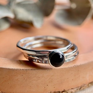 May include: Three silver rings with a black gemstone in the centre ring. The rings are stacked on top of each other and are on a light brown surface.