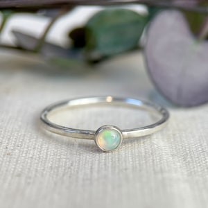 May include: A delicate silver ring featuring a small, round opal gemstone. The opal has a milky white colour with hints of green and blue.