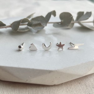 May include: Five silver stud earrings with different shapes: a circle, a heart, a crescent moon, a star, and a lightning bolt.