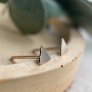 May include: A pair of gold triangle stud earrings on a light brown surface.