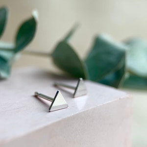 May include: Two silver triangle stud earrings on a light pink surface. The earrings are small and delicate.