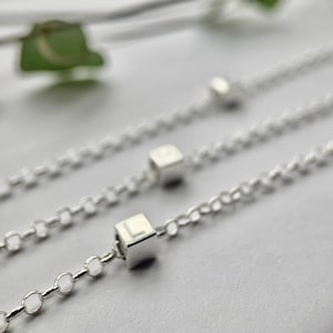 May include: Three silver chains with small silver cube charms. Each charm has a letter engraved on it. The letter on the charm in focus is "L".