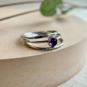 May include: Three silver rings stacked on top of each other. The top ring has a small, round, purple amethyst gemstone.