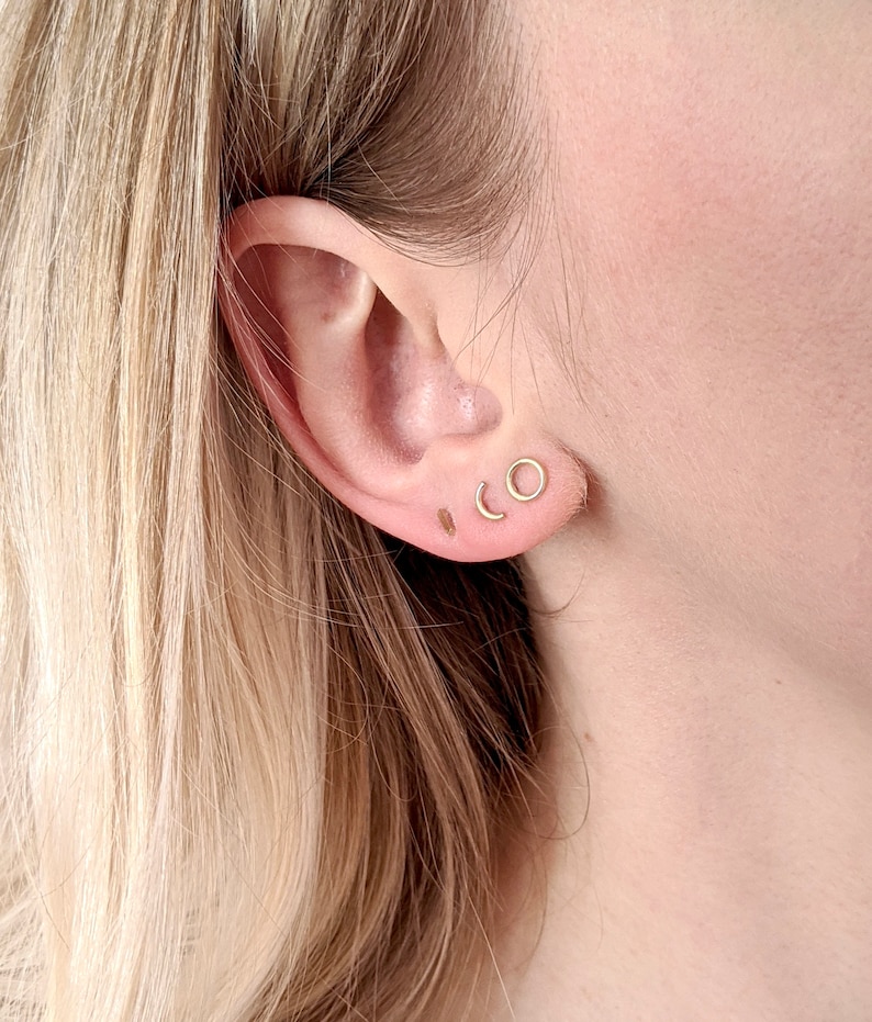 May include: A close-up of a person's ear with three small gold earrings. The earrings are a circle, a crescent moon, and a thin bar.