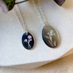 May include: Two silver necklaces with oval pendants featuring engraved floral designs. One pendant has a delicate flower design, while the other features a larger, more intricate flower.