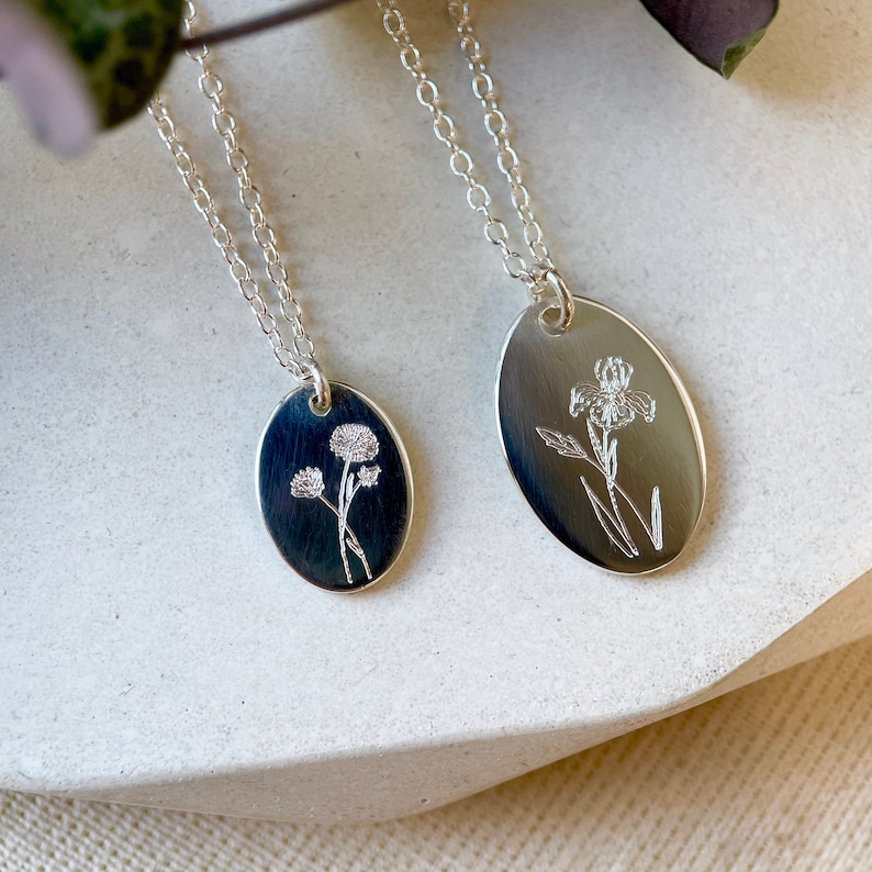 May include: Two silver necklaces with oval pendants featuring engraved floral designs. One pendant has a delicate flower design, while the other features a larger, more intricate flower.