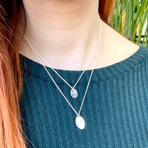 May include: Two silver necklaces with delicate chains and small oval pendants. The pendants feature a simple floral design.