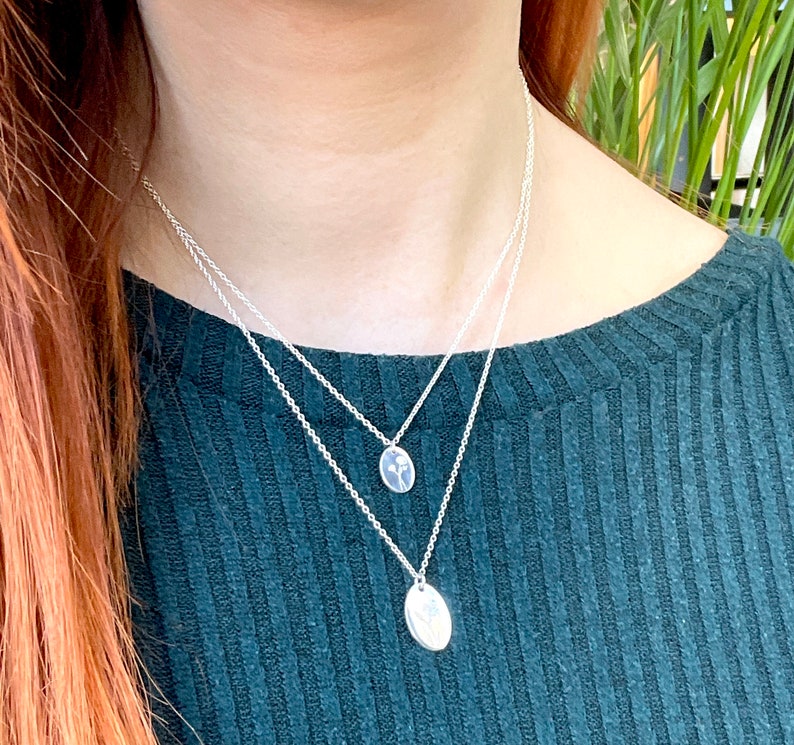 May include: Two silver necklaces with delicate chains and small oval pendants. The pendants feature a simple floral design.