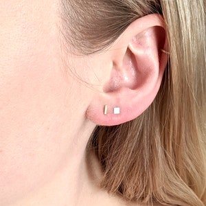 May include: A close-up of a person's ear with two small gold earrings. One earring is a square shape and the other is a thin rectangle.