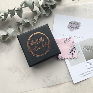 May include: A black jewelry box with a gold foil logo that reads "the little silver kile". The box is sitting on a white surface with a pink and white care instruction card and a thank you card.