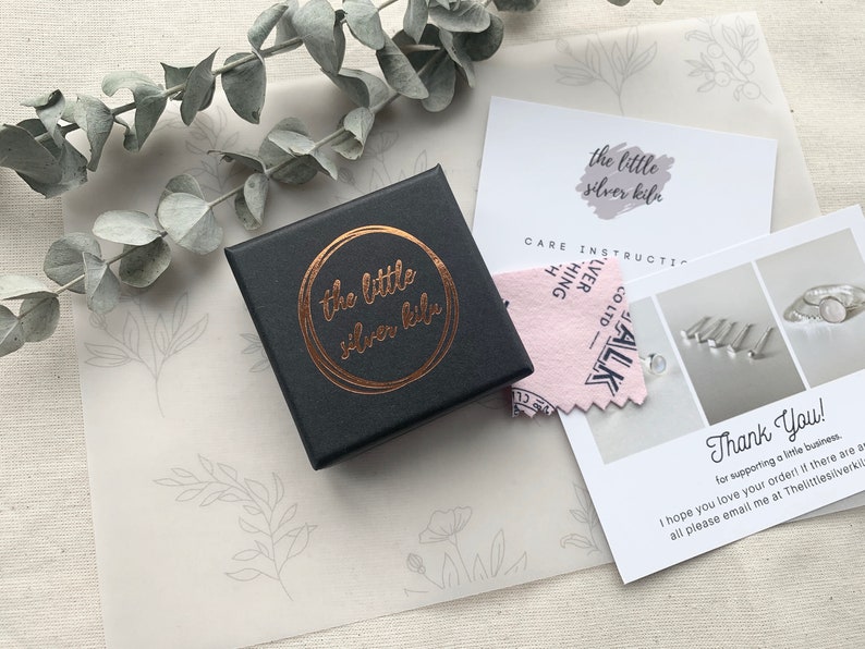 May include: A black jewelry box with a gold foil logo that reads "the little silver kile". The box is sitting on a white surface with a pink and white care instruction card and a thank you card.