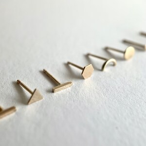 May include: A set of seven minimalist gold earrings with different geometric shapes, including a triangle, rectangle, hexagon, crescent moon, and circle.