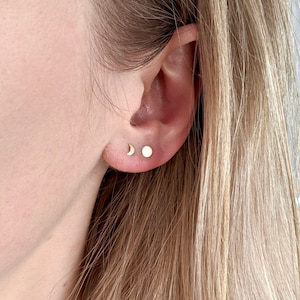 May include: A close-up of a person's ear with two small gold earrings. The earrings are a crescent moon and a small circle.