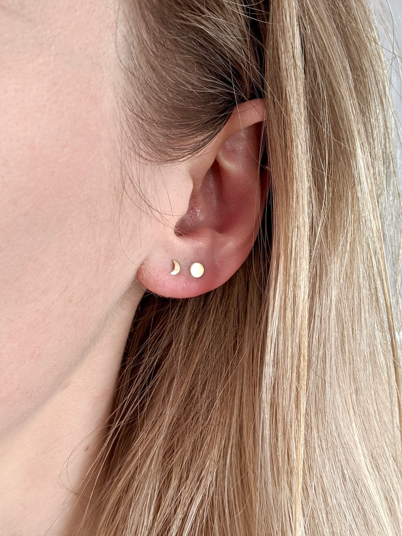 May include: A close-up of a person's ear with two small gold earrings. The earrings are a crescent moon and a small circle.