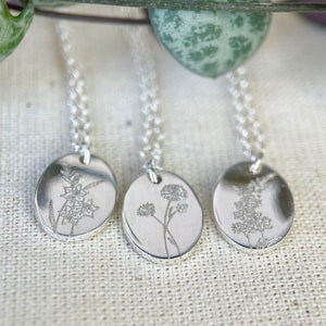 May include: Three silver necklaces with delicate floral designs on oval pendants. The necklaces are on a white fabric background.