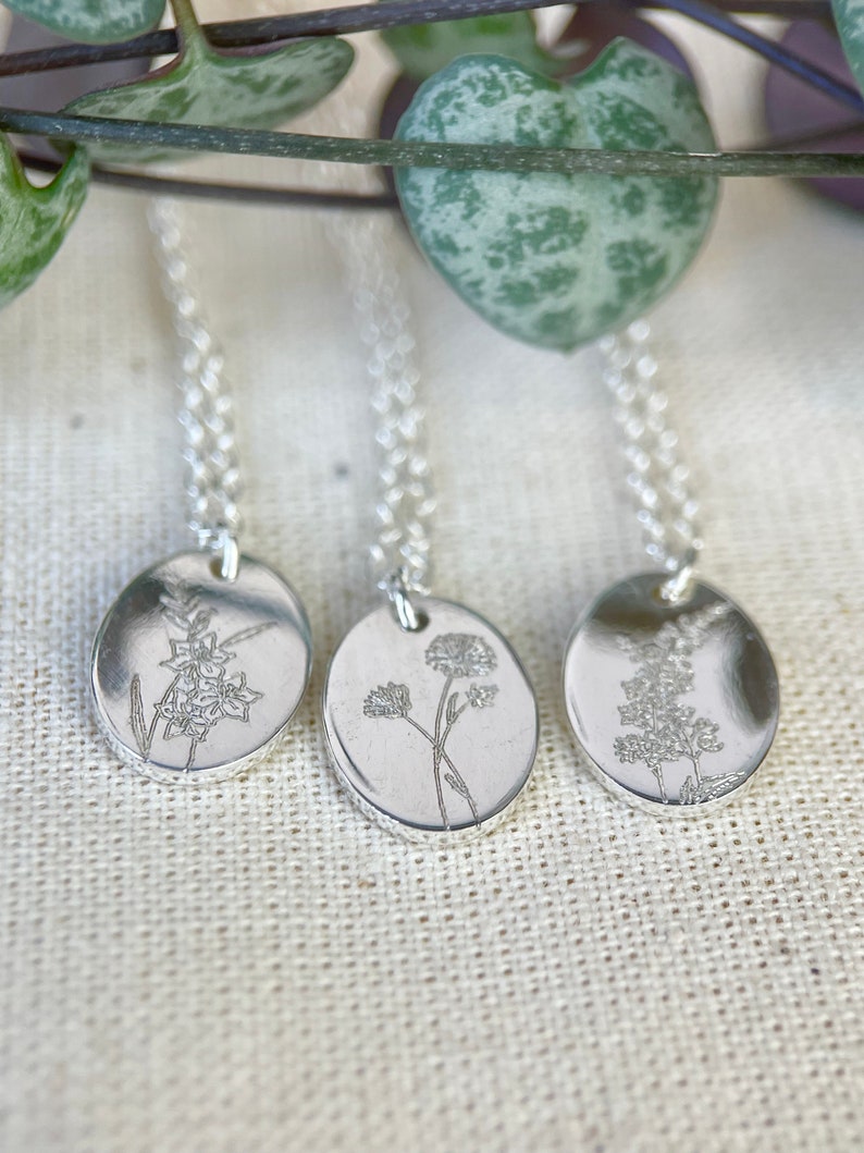 May include: Three silver necklaces with delicate floral designs on oval pendants. The necklaces are on a white fabric background.
