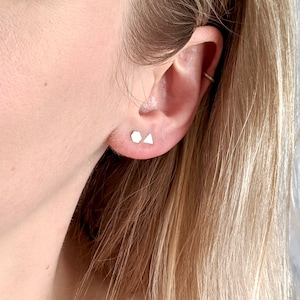 May include: A close-up of a person's ear with two small, silver geometric earrings. The earrings are in the shape of a triangle and a square.