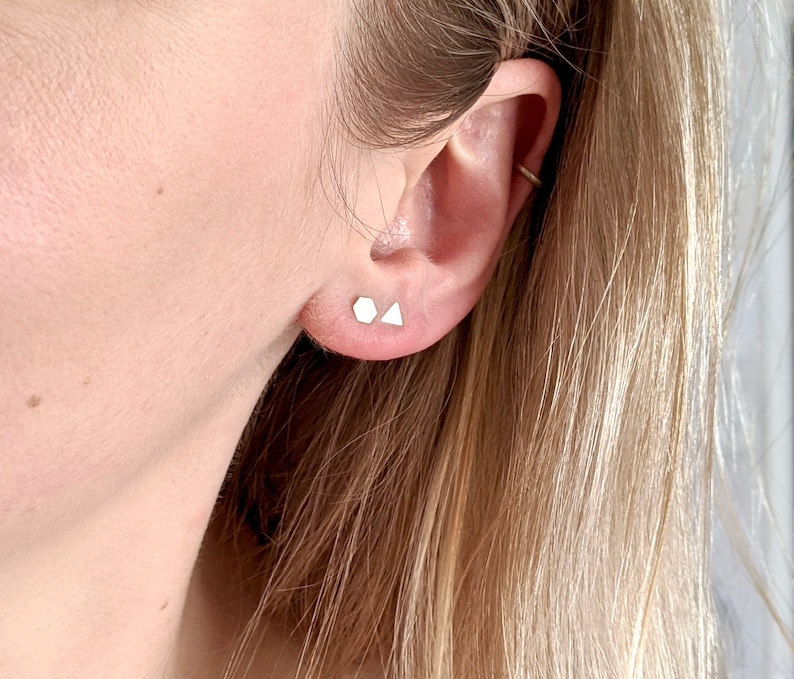 May include: A close-up of a person's ear with two small, silver geometric earrings. The earrings are in the shape of a triangle and a square.
