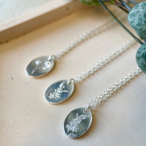 May include: Three silver necklaces with oval pendants featuring delicate pressed flower designs. The necklaces are displayed on a light beige surface.