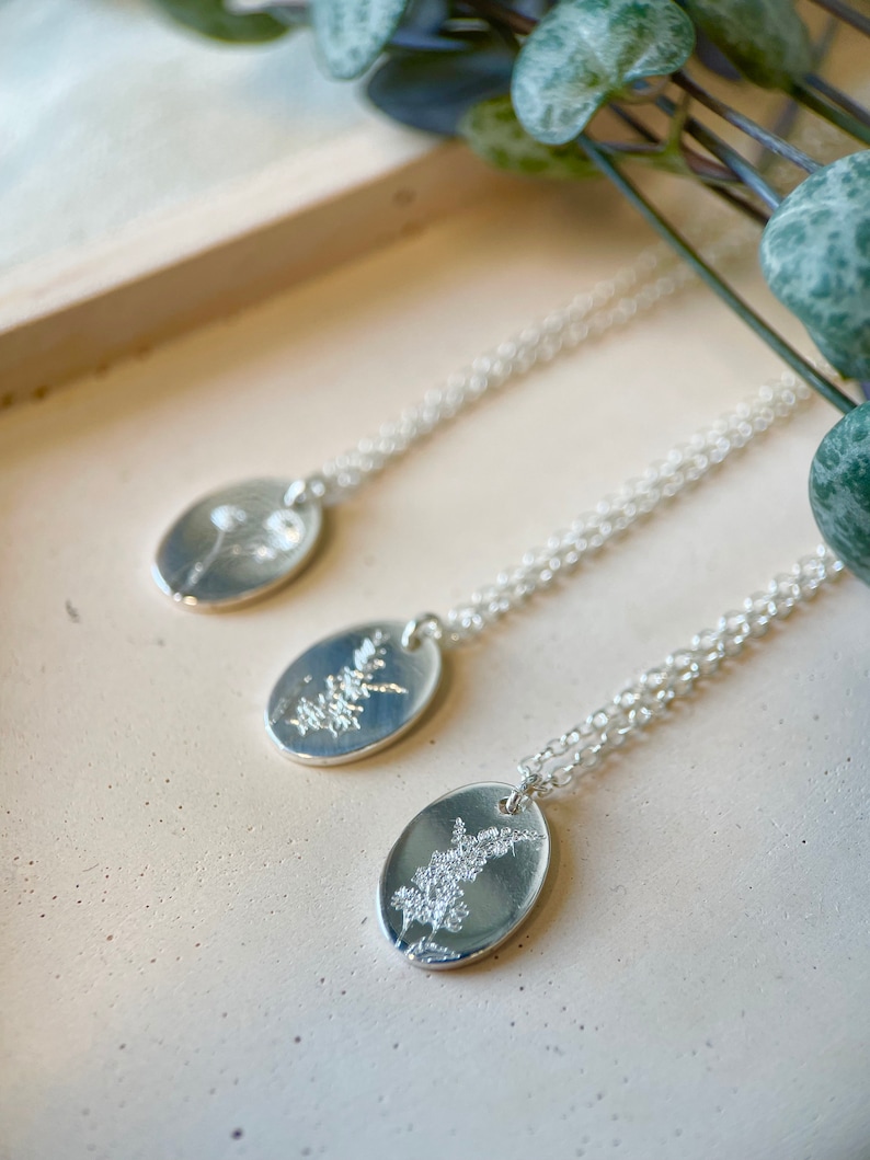 May include: Three silver necklaces with oval pendants featuring delicate pressed flower designs. The necklaces are displayed on a light beige surface.