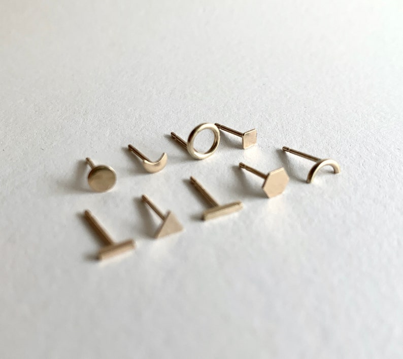 May include: A set of nine gold-toned stud earrings featuring different geometric shapes, including a circle, square, hexagon, crescent moon, triangle, and a line.