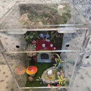 Puede incluir: Un terrario de acr&iacute;lico transparente con un jard&iacute;n de hadas en miniatura en su interior. El jard&iacute;n presenta un estanque con peces, una casa de setas y varias plantas y setas. El terrario es hexagonal y tiene una tapa con bisagras.