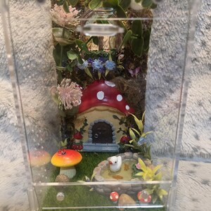 Puede incluir: Un terrario en miniatura con una escena de jard&iacute;n de hadas caprichosa. Presenta una casa de hongo roja y blanca, un estanque con un pato blanco y flores coloridas. El recinto de acr&iacute;lico transparente muestra el detallado paisaje en miniatura.