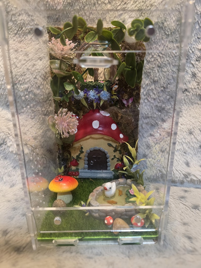 Puede incluir: Un terrario en miniatura con una escena de jard&iacute;n de hadas caprichosa. Presenta una casa de hongo roja y blanca, un estanque con un pato blanco y flores coloridas. El recinto de acr&iacute;lico transparente muestra el detallado paisaje en miniatura.
